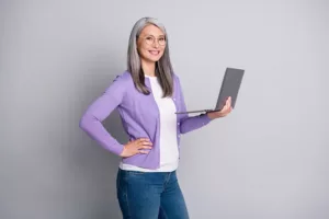 Photo portrait of smiling friendly elder female coach keeping laptop posing wearing transparent eyeglasses isolated on grey color background